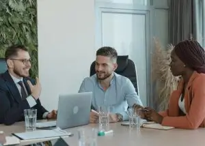 Tre professionisti in una riunione, conversano sorridendo con un laptop sul tavolo, simbolo di crescita professionale e collaborazione